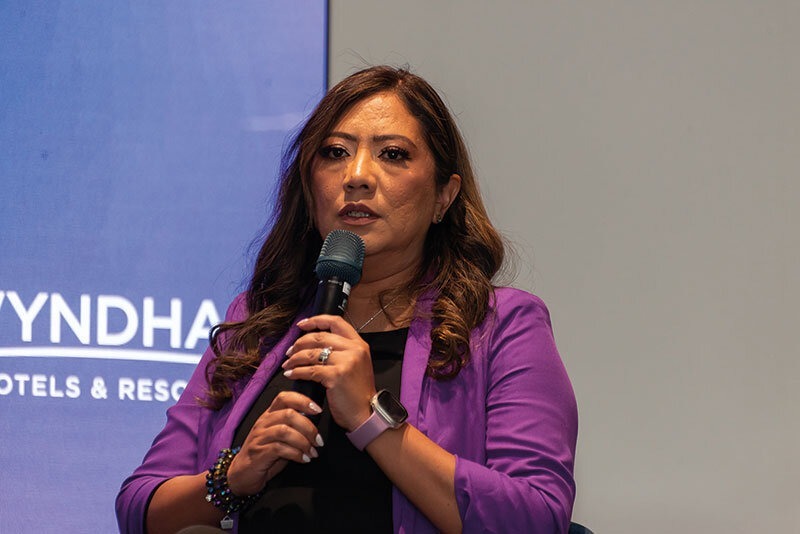 Una conferencista de alto perfil, reconocida por su experiencia en el sector inmobiliario de lujo, exponente sobre innovación tecnológica en bienes raíces (proptech). Vestida elegantemente con un blazer púrpura, comparte sus conocimientos y perspectivas ante una audiencia selecta e inversores exigentes. El evento se desarrolla en un entorno corporativo exclusivo, acompañado del emblema de Wyndham Hotels & Resorts como referente global de excelencia y prestigio en hospitalidad.