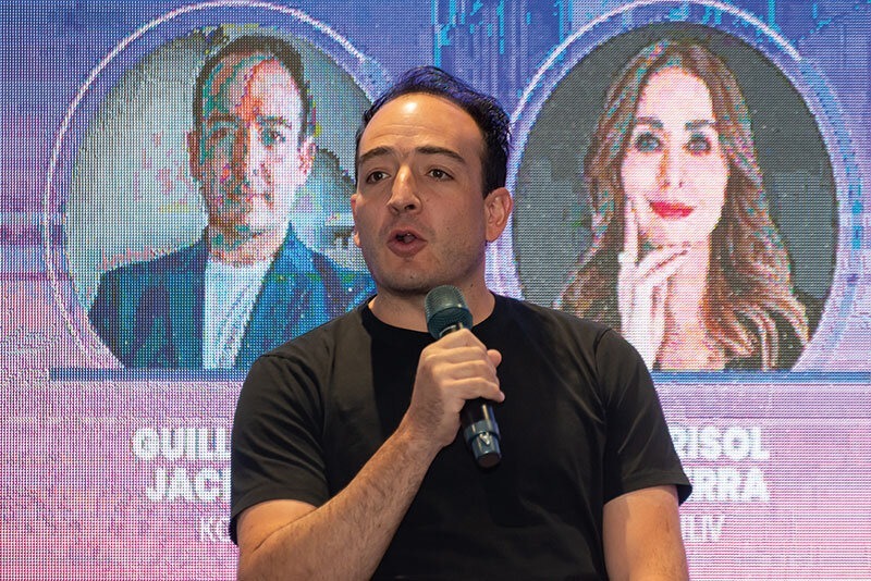 Un profesional del sector inmobiliario, vestido de manera sobria, interviene como ponente en una conferencia especializada sobre proptech. Mientras exponen su visión innovadora ante una audiencia de alto nivel, dos proyecciones a gran escala presentan a destacados referentes del sector, resaltando su experticia y protagonismo en la transformación digital del mercado inmobiliario premium. Esta escena refleja el liderazgo intelectual y la apuesta por tecnología de vanguardia que definen las tendencias más exclusivas para inversores y compradores exigentes.