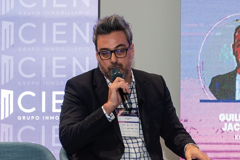 Un ejecutivo de amplia trayectoria en el sector inmobiliario premium, distinguido por su cabello canoso y gafas, viste un blazer negro y una camisa a cuadros mientras interviene como ponente ante un público selecto. Porta una credencial oficial, reflejo de su participación en un evento exclusivo organizado por CIEN Grupo Inmobiliario. Al fondo, se exhibe un banner corporativo que resalta los avances más innovadores en proptech e impulsa la visión de vanguardia del grupo; junto a este, una pantalla proyecta información relevante para inversores y compradores exigentes interesados en oportunidades diferenciadas dentro del mercado inmobiliario de alto valor.