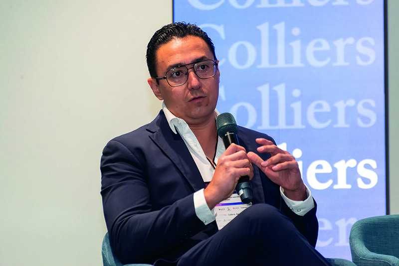 Un distinguido ponente, ataviado con un elegante traje azul marino y gafas, exponen sobre temas de geopolítica durante un evento exclusivo del sector inmobiliario. Su credencial de identificación resalta su participación como orador principal. El escenario cuenta con una sofisticada señalización corporativa en azul, destacando la marca Colliers, símbolo de excelencia y liderazgo en el mercado inmobiliario premium.