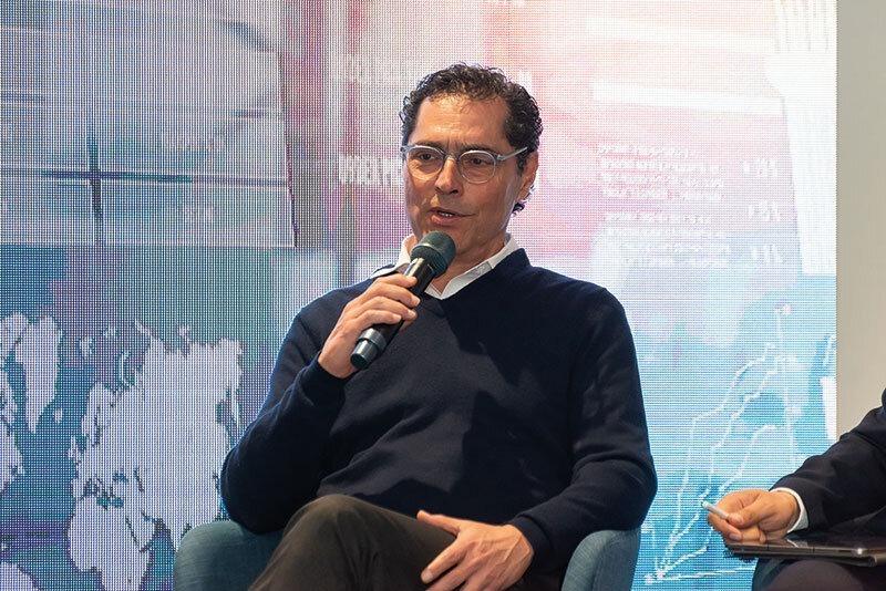 Un distinguido ponente, reconocido por su experiencia en geopolítica, participa como conferenciante principal en un evento exclusivo. Ataviado con indumentaria elegante y moderna, sostiene el micrófono mientras comparte perspectivas estratégicas ante una audiencia selecta. El fondo, compuesto por un mapa mundial artístico y elementos abstractos de diseño contemporáneo, refuerza la atmósfera global e innovadora del encuentro. Esta escena ejemplifica el nivel de sofisticación y visión internacional que caracteriza a las experiencias asociadas con los activos inmobiliarios premium: espacios donde convergen líderes influyentes para debatir tendencias que impactan el mercado mundial y generan oportunidades de inversión excepcionales.