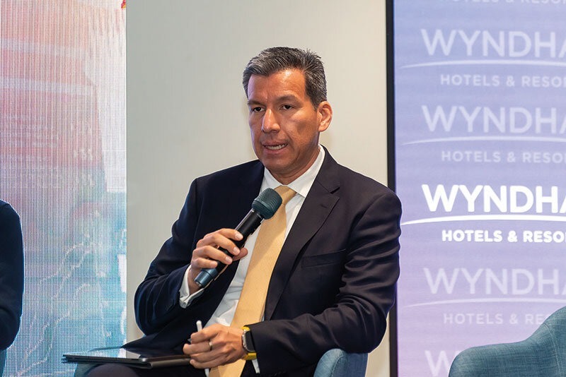 Un ejecutivo de alto nivel, vestido con un elegante traje oscuro y corbata amarilla, se dirige a una audiencia especializada mientras analiza temas de geopolítica con visión estratégica. Sentado con porte profesional, sostiene un micrófono en la mano y toma notas cuidadosamente, demostrando su enfoque analítico. De fondo sobresale el emblemático logo de Wyndham Hotels & Resorts, sinónimo de excelencia y liderazgo global en hospitalidad premium, un contexto ideal para inversores y compradores que buscan oportunidades exclusivas en el mercado inmobiliario internacional.