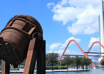 Una imponente escultura industrial de tono óxido se erige sobre una plataforma junto al canal, en una de las ubicaciones más privilegiadas de Nuevo León. Al fondo, la silueta vanguardista de un rascacielos blanco y la arquitectura singular de un puente naranja en arco subrayan el dinamismo urbano y el desarrollo sofisticado que caracterizan a este enclave. Todo ello, enmarcado por un cielo azul con nubes dispersas, ofrece la perspectiva ideal para inversionistas y compradores que buscan integración única entre arte contemporáneo, infraestructura modernista y exclusividad inmobiliaria.