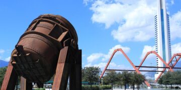 Una imponente escultura industrial de tono óxido se erige sobre una plataforma junto al canal, en una de las ubicaciones más privilegiadas de Nuevo León. Al fondo, la silueta vanguardista de un rascacielos blanco y la arquitectura singular de un puente naranja en arco subrayan el dinamismo urbano y el desarrollo sofisticado que caracterizan a este enclave. Todo ello, enmarcado por un cielo azul con nubes dispersas, ofrece la perspectiva ideal para inversionistas y compradores que buscan integración única entre arte contemporáneo, infraestructura modernista y exclusividad inmobiliaria.