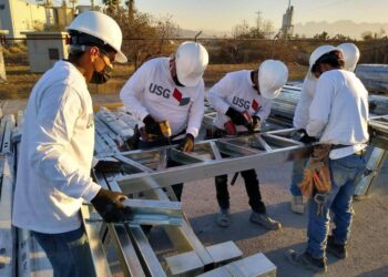 Un equipo de cinco profesionales altamente capacitados, equipados con cascos blancos y guantes de seguridad USG, trabaja en la instalación de estructuras metálicas en un entorno industrial. Su vestimenta incluye camisetas corporativas con el logotipo de USG, reflejando los más altos estándares del sector. De fondo, se aprecian edificaciones industriales que realzan la solidez e innovación del proyecto en desarrollo. Esta escena destaca el compromiso con la calidad constructiva y la excelencia operativa, cualidades clave para inversores y compradores que buscan activos premium en el mercado inmobiliario.