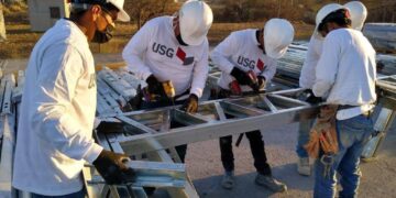 Un equipo de cinco profesionales altamente capacitados, equipados con cascos blancos y guantes de seguridad USG, trabaja en la instalación de estructuras metálicas en un entorno industrial. Su vestimenta incluye camisetas corporativas con el logotipo de USG, reflejando los más altos estándares del sector. De fondo, se aprecian edificaciones industriales que realzan la solidez e innovación del proyecto en desarrollo. Esta escena destaca el compromiso con la calidad constructiva y la excelencia operativa, cualidades clave para inversores y compradores que buscan activos premium en el mercado inmobiliario.
