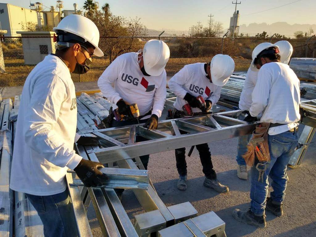 Un equipo de cinco profesionales altamente capacitados, equipados con cascos blancos y guantes de seguridad USG, trabaja en la instalación de estructuras metálicas en un entorno industrial. Su vestimenta incluye camisetas corporativas con el logotipo de USG, reflejando los más altos estándares del sector. De fondo, se aprecian edificaciones industriales que realzan la solidez e innovación del proyecto en desarrollo. Esta escena destaca el compromiso con la calidad constructiva y la excelencia operativa, cualidades clave para inversores y compradores que buscan activos premium en el mercado inmobiliario.