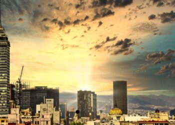 Panorámica urbana al atardecer que revela un horizonte cómodo, dominado por imponentes rascacielos y una torre emblemática al lado izquierdo. El horizonte, enmarcado por nubes dispersas y la luz dorada del sol filtrándose entre dos edificios principales, resalta el dinamismo y la exclusividad de una ciudad cosmopolita ideal para inversiones inmobiliarias premium.