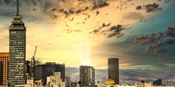 Panorámica urbana al atardecer que revela un horizonte cómodo, dominado por imponentes rascacielos y una torre emblemática al lado izquierdo. El horizonte, enmarcado por nubes dispersas y la luz dorada del sol filtrándose entre dos edificios principales, resalta el dinamismo y la exclusividad de una ciudad cosmopolita ideal para inversiones inmobiliarias premium.