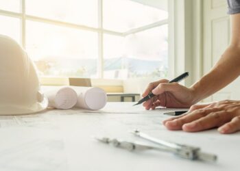 Un profesional revisa detalladamente planos arquitectónicos sobre su escritorio, utilizando instrumentos de precisión como un compás y una pluma. En la superficie destacan un casco de seguridad blanco y planos enrollados, reflejando el compromiso con los más altos estándares en diseño y construcción premium. La abundante luz natural que ingresa por los amplios ventanales acentúa la modernidad y el enfoque en espacios luminosos propios del sector inmobiliario de alto nivel. Escenarios como este son características en proyectos distinguidos, posibles candidatos a reconocimientos internacionales como los premios IWBI 2024, donde la excelencia en innovación y sostenibilidad es prioridad para inversionistas y compradores exigentes.