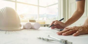 Un profesional revisa detalladamente planos arquitectónicos sobre su escritorio, utilizando instrumentos de precisión como un compás y una pluma. En la superficie destacan un casco de seguridad blanco y planos enrollados, reflejando el compromiso con los más altos estándares en diseño y construcción premium. La abundante luz natural que ingresa por los amplios ventanales acentúa la modernidad y el enfoque en espacios luminosos propios del sector inmobiliario de alto nivel. Escenarios como este son características en proyectos distinguidos, posibles candidatos a reconocimientos internacionales como los premios IWBI 2024, donde la excelencia en innovación y sostenibilidad es prioridad para inversionistas y compradores exigentes.