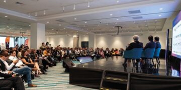 Un panel de expertos inmobiliarios se encuentra en el escenario, dirigiéndose a una audiencia selecta en un cómodo salón de conferencias. Los asistentes, atentos y comprometidos—muchos tomando notas y capturando imágenes clave—demuestran un interés genuino por las tendencias más exclusivas del sector. Una pantalla de gran formato acompaña la presentación, ofreciendo información relevante y actualizada sobre oportunidades de inversión premium, dinámicas del mercado y desarrollos emblemáticos para compradores e inversores que buscan excelencia y valor agregado en bienes raíces de alto nivel.