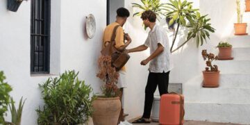 Dos personas se encuentran frente a una exclusiva fachada blanca, embellecida con elegantes plantas en maceta. Una de ellas accede al interior utilizando una llave y transportando una maleta de viaje, evocando la sofisticación y hospitalidad propia de los desarrollos inmobiliarios premium. La otra, con bolso al hombro, complementa este entorno distinguido que responde a las expectativas de compradores e inversores que valoran el diseño superior y la experiencia residencial de alto nivel.