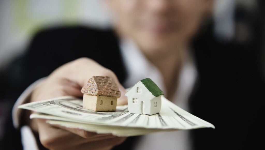 Una persona sostiene dos exclusivos modelos de inmuebles y un fajo de billetes estadounidenses, simbolizando sólidas oportunidades de inversión en el sector inmobiliario premium. La imagen enfatiza el valor tangible y el potencial de alta rentabilidad de los activos, mientras que la figura humana, deliberadamente desenfocada en segundo plano, subraya la importancia del patrimonio sobre lo individual. Se presenta así una poderosa representación visual del proceso estratégico de recuperación y gestión eficiente de carteras inmobiliarias selectas para inversores y compradores exigentes.