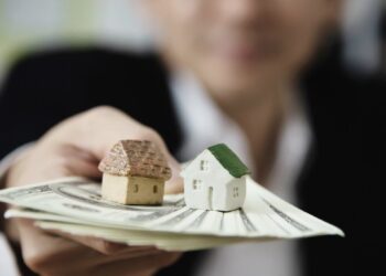 Una persona sostiene dos exclusivos modelos de inmuebles y un fajo de billetes estadounidenses, simbolizando sólidas oportunidades de inversión en el sector inmobiliario premium. La imagen enfatiza el valor tangible y el potencial de alta rentabilidad de los activos, mientras que la figura humana, deliberadamente desenfocada en segundo plano, subraya la importancia del patrimonio sobre lo individual. Se presenta así una poderosa representación visual del proceso estratégico de recuperación y gestión eficiente de carteras inmobiliarias selectas para inversores y compradores exigentes.