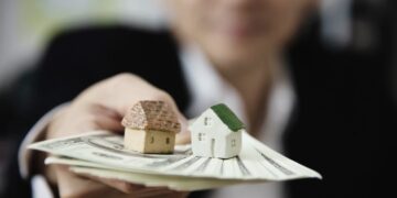 Una persona sostiene dos exclusivos modelos de inmuebles y un fajo de billetes estadounidenses, simbolizando sólidas oportunidades de inversión en el sector inmobiliario premium. La imagen enfatiza el valor tangible y el potencial de alta rentabilidad de los activos, mientras que la figura humana, deliberadamente desenfocada en segundo plano, subraya la importancia del patrimonio sobre lo individual. Se presenta así una poderosa representación visual del proceso estratégico de recuperación y gestión eficiente de carteras inmobiliarias selectas para inversores y compradores exigentes.