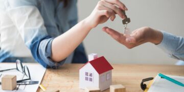 Una persona entrega la llave de una propiedad a otra, sobre una mesa con documentación, una maqueta residencial y bloques de madera. Esta escena representa el dinámico mercado inmobiliario premium en México, donde la activa demanda por inmuebles de alta gama evidencia el atractivo y solidez del sector para inversores y compradores beneficiosos que buscan activos exclusivos con alto potencial de valorización.