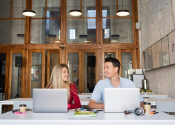 Dos profesionales ocupan una mesa en oficinas de diseño contemporáneo, caracterizadas por ventanas de gran altura y elegantes marcos de madera. El ambiente, cuidadosamente concebido para potenciar la productividad y fomentar la colaboración, representa un entorno ideal para inversores y compradores que valoran espacios empresariales exclusivos, funcionales y a la vanguardia del sector inmobiliario premium.