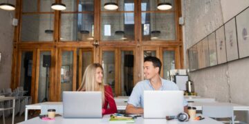 Dos profesionales ocupan una mesa en oficinas de diseño contemporáneo, caracterizadas por ventanas de gran altura y elegantes marcos de madera. El ambiente, cuidadosamente concebido para potenciar la productividad y fomentar la colaboración, representa un entorno ideal para inversores y compradores que valoran espacios empresariales exclusivos, funcionales y a la vanguardia del sector inmobiliario premium.