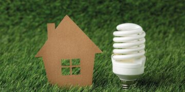 Un recorte representativo de una vivienda y una lámpara de bajo consumo se sitúan sobre césped natural, evocando el concepto de vivienda sustentable. Esta imagen transmite un firme compromiso con la eficiencia energética y la responsabilidad ambiental, elementos esenciales en el segmento inmobiliario premium para inversores y compradores que priorizan la innovación, la sostenibilidad y el valor a largo plazo.