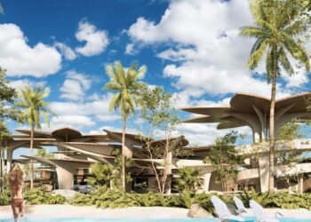 Dos personas se acercan a una exclusiva piscina rodeada de exuberante vegetación tropical, creando un entorno de privacidad y sofisticación absoluta. Al fondo, se alza un moderno edificio Muúk Karant cuya arquitectura vanguardista destaca por sus imponentes techos en forma de hoja, integrándose armónicamente con el paisaje natural. Bajo un cielo azul salpicado de nubes, esta propiedad representa una oportunidad incomparable para inversores y compradores que buscan excelencia estética, innovación arquitectónica y contacto directo con la naturaleza en el segmento inmobiliario premium.