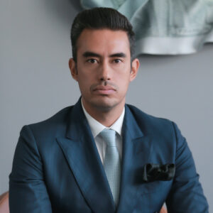 Un hombre vestido con un traje azul, camisa blanca y corbata gris claro se presenta con una expresión serena en One Laguna Cancún, el desarrollo inmobiliario premium de referencia. La elegancia de los detalles-como el pañuelo negro en su chaqueta-refleja el nivel de sofisticación que caracteriza a este exclusivo entorno. Al fondo, discretos elementos en tonos grises complementan la atmósfera contemporánea y refinada del proyecto, ideal para inversionistas y compradores que buscan distinción, plusvalía y calidad inigualable.