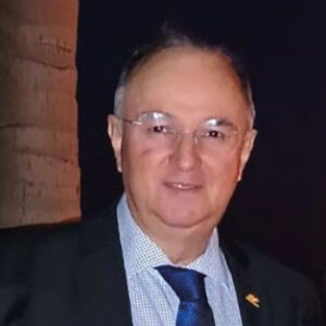 Ejemplo reescrito con tono profesional y aspiracional, dirigido al sector inmobiliario premium: Un ejecutivo de alto nivel, con traje oscuro, camisa a cuadros en tonos blanco y azul y corbata azul, se presenta ante un fondo sobrio realzado por una imponente columna de piedra. Su presencia liderazgo transmite y dominio en el financiamiento de proyectos inmobiliarios exclusivos, atributos esenciales para inversores y compradores que buscan oportunidades excepcionales en el mercado de lujo.