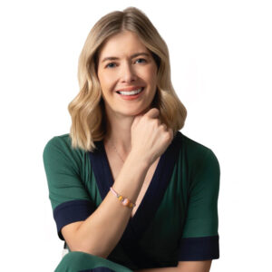 Una mujer de cabello rubio, vestida con un conjunto en tonos verdes y azul marino, posa sonriente en un entorno neutro. Su postura, apoyando la mano bajo el mentón y luciendo una elegante pulsera de cuentas coloridas, refleja confianza y determinación: cualidades esenciales para afrontar retos y encontrar soluciones efectivas. Esta imagen transmite el compromiso profesional e inspirador necesario en el sector inmobiliario premium, orientado a quienes valoran la excelencia y buscan oportunidades exclusivas de inversión o adquisición.