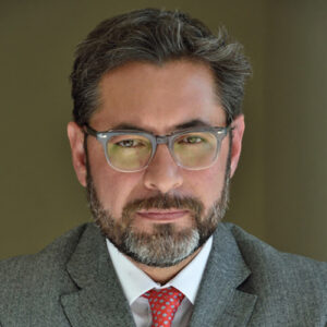 Distinguido profesional de cabello canoso y barba bien cuidada, con gafas y atuendo impecable—traje gris, camisa blanca y corbata roja con detalles—mira directamente a la cámara desde un fondo neutro. Su presencia transmite autoridad y experiencia en Building Science y desarrollo inmobiliario de alto nivel, atributos esenciales para liderar proyectos premium orientados a inversores exigentes que buscan excelencia, innovación y valor sostenible en el sector inmobiliario.