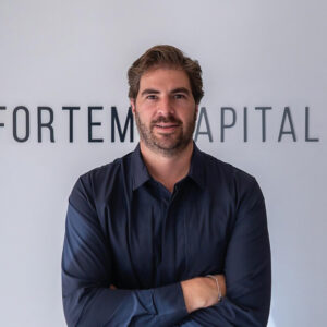 Un hombre de porte seguro, con cabello castaño medio y barba bien cuidada, posa con los brazos cruzados frente a una pared en la que se distingue parcialmente el nombre FORTEM CAPITAL. Esta firma es reconocida por su experiencia en el financiamiento de proyectos inmobiliarios de alto nivel. Vestido con camisa oscura, proyecta confianza y profesionalismo, valores clave para inversores y compradores que buscan excelencia y solidez en el sector premium.