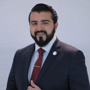 Un hombre de cabello oscuro y barba perfectamente cuidada, vestido con un elegante traje oscuro, camisa blanca impecable y corbata roja, se presenta sobre un fondo claro y sobrio. Su leve sonrisa transmite seguridad y profesionalismo, cualidades esenciales en los líderes del financiamiento inmobiliario premium. La imagen proyecta confianza y solvencia, atributos altamente valorados por inversores y compradores que buscan excelencia en cada etapa de sus operaciones inmobiliarias.