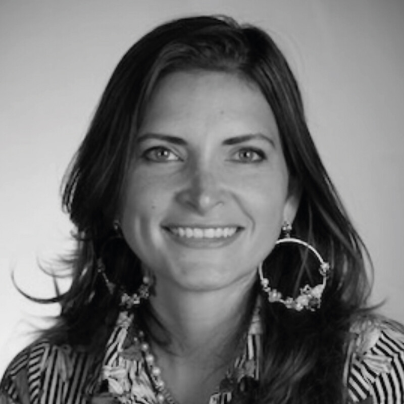 Imagen en blanco y negro de una mujer con larga cabellera oscura, que transmite confianza y sofisticación a través de su sonrisa. Luce pendientes circulares de gran tamaño y una blusa de diseño exclusivo inspirado en el estilo distintivo de Baja. El fondo, neutro y desenfocado, dirige toda la atención hacia la protagonista, resaltando su presencia magnética—un reflejo del carácter único que distingue a quienes buscan propiedades premium en destinos selectos.