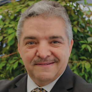 Un profesional de amplia trayectoria en Ciencias de la Construcción, con cabello canoso y bigote, viste un elegante traje negro y corbata con diseño distintivo. Su actitud cordial y segura refleja experiencia, solvencia y liderazgo en el sector inmobiliario premium. La imagen se toma en un entorno natural, que aporta exclusividad al retrato; el especialista representa los más altos estándares de calidad e innovación para inversores y compradores exigentes.