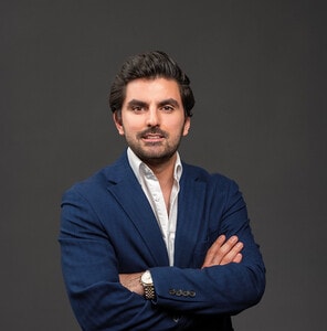 Varón de porte distinguido, cabello oscuro y barba cuidada, posa frente a un fondo sobrio y elegante. Viste un impecable saco azul marino sobre camisa blanca, símbolo de profesionalismo y sofisticación. Con brazos cruzados y expresión serena pero segura, se presenta como un asesor experto dispuesto a analizar las mejores oportunidades de inversión en el sector inmobiliario premium, especialmente ante el dinámico panorama que ofrece el nearshoring para inversores visionarios.