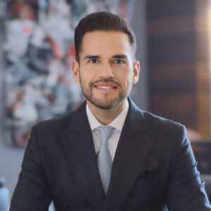 Un hombre de cabello oscuro y barba perfectamente recortada luce un elegante traje oscuro, camisa blanca y corbata en tono claro. Se muestra sonriente mientras posa en el interior de One Laguna Cancún, con un fondo abstracto que evoca la exclusividad y sofisticación del Caribe mexicano. Este entorno refleja el estilo contemporáneo y la atención al detalle que caracteriza a las propiedades premium, ideales para inversionistas y compradores que valoran el lujo, la ubicación privilegiada y una experiencia residencial inigualable.