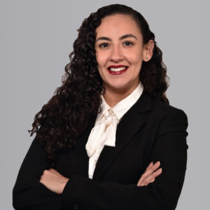 Una profesional de presencia impecable, distinguida por su cabello oscuro y rizado, viste un elegante conjunto de blazer negro y blusa blanca con lazo. De pie frente a un fondo gris sobrio y moderno, irradia seguridad mientras posa con los brazos cruzados y una expresión confiada. Su liderazgo se refleja en su capacidad para abordar con autoridad los retos estratégicos asociados al nearshoring, cualidad clave para quienes buscan asesoría experta en inversiones inmobiliarias premium que demandan excelencia, visión internacional y minimización efectiva del riesgo.