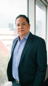 Un hombre viste un elegante saco oscuro y camisa azul a cuadros mientras posa junto a un ventanal de gran formato, con vistas panorámicas de la ciudad y áreas arboladas al fondo. Su puerta transmite la seguridad característica de los principales actores en el análisis del mercado inmobiliario premium en México. De apariencia sereno y profesional, personifica la confianza y visión estratégica que buscan los inversores más exigentes del sector.