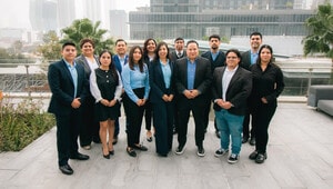 Un distinguido grupo de trece profesionales, vestidos con elegancia en trajes formales y de negocios, posa en dos filas sobre una exclusiva terraza pavimentada. De fondo, se aprecian edificios contemporáneos perfectamente integrados con áreas verdes, creando un entorno cómodo que refleja modernidad y alto valor patrimonial. Este equipo representa el más alto nivel de experticia en inteligencia estratégica aplicada al sector industrial premium en México, siendo la elección preferente para inversionistas y compradores que buscan activos inmobiliarios excepcionales y oportunidades sólidas de crecimiento.