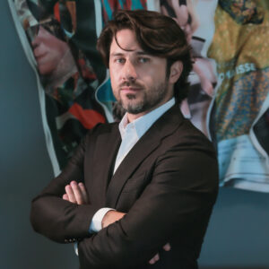 Un hombre de cabello oscuro y barba bien cuidada, vestido con un elegante traje en tonos oscuros y camisa blanca impecable, posa con los brazos cruzados frente a un fondo abstracto que evoca la energía vibrante del Caribe mexicano y la exclusividad de Laguna Cancún. Esta imagen transmite confianza, sofisticación y una visión alineada con el estándar de los inversionistas y compradores más exigentes del sector inmobiliario premium.