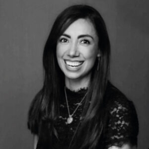 Una mujer de cabello oscuro y largo, ataviada con una blusa de encaje y delicados collares, posa sonriente ante un fondo neutro. Esta imagen en blanco y negro transmite sofisticación y distinción: atributos que reflejan a la perfección el estilo de vida exclusivo que define al sector inmobiliario premium en Los Cabos. Una invitación visual para quienes buscan inversiones excepcionales en destinos reconocidos por su elegancia y atractivo internacional.