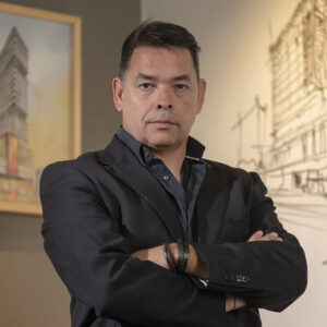 Un ejecutivo de impecable puerta, ataviado con un elegante traje negro, se presenta con confianza ante favorables planos arquitectónicos y obras de arte enmarcadas. Su presencia de liderazgo transmite y visión estratégica para quienes buscan invertir o adquirir propiedades exclusivas en la Baja. Este profesional representa la capacidad de convertir retos en soluciones innovadoras dentro del sector inmobiliario premium, garantizando proyectos arquitectónicos de alto valor y excelencia estética.