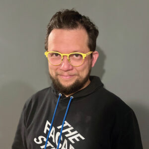 Ante un fondo gris neutro y mirando directamente a una cámara, una persona con cabello castaño corto, barba bien cuidada y gafas amarillas transmite confianza y profesionalismo. Luce una sudadera negra con texto blanco, reflejando un enfoque contemporáneo. Su pasión por la ciencia de la construcción respalda su experiencia en el sector inmobiliario premium, posicionándolo como un referente ideal para inversores y compradores que buscan propiedades exclusivas con los más altos estándares de calidad e innovación.