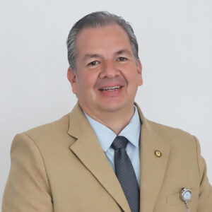 Un distinguido profesional del sector inmobiliario premium, con impecable porte y vestimenta ejecutiva en tonos beige y azul, posa con una actitud segura frente a un fondo minimalista. Su cabello entrecano refleja experiencia, mientras una insignia en su solapa destaca su participación activa en la búsqueda de soluciones innovadoras e inteligentes ante los retos arquitectónicos más exigentes de Baja California. Esta sólida combinación de experiencia, visión y estratégica lo posiciona como un aliado para inversionistas o compradores que buscan proyectos exclusivos con altos estándares de calidad y rendimiento.