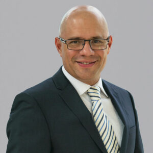 Un ejecutivo de alto nivel, vestido con un elegante traje oscuro, camisa blanca impecable y corbata a rayas, posa ante un fondo gris claro. Su expresión serena y segura transmite la confianza esencial para liderar iniciativas en el sector inmobiliario premium o gestionar estrategias avanzadas de financiamiento de proyectos. Un perfil idóneo para quienes buscan asociarse con profesionales capaces de impulsar inversiones sólidas y exitosas.