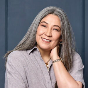 Una mujer de cabello canoso y largo, vestida con una camisa abotonada en tono lavanda, posa con elegancia y confianza frente a la cámara. Luce un reloj dorado y pendientes tipo aro, proyectando sofisticación y profesionalismo sobre un fondo azul oscuro. Esta imagen transmite la experiencia, serenidad y liderazgo propios de una ejecutiva especializada en edificaciones sustentables—un retrato ideal para perfiles profesionales orientados al sector inmobiliario premium o inversiones certificadas LEED.
