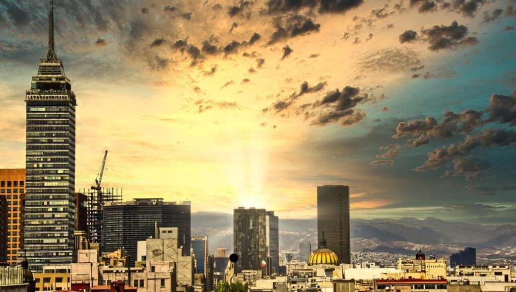 El horizonte urbano se presenta como un sofisticado equilibrio entre arquitectura contemporánea y patrimonio histórico, coronado al atardecer por la silueta imponente de un rascacielos emblemático en primer plano. Destaca la icónica estructura de cúpula dorada, símbolo del dinamismo y crecimiento sostenido de las FIBRAs en México, perfilándose como epicentro de las inversiones inmobiliarias premium. El entorno se realza con una vista majestuosa hacia las montañas y un cielo cautivador, evocando exclusividad y potencial de plusvalía para compradores e inversionistas que exigen excelencia arquitectónica y ubicaciones privilegiadas.