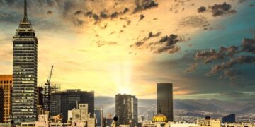 El horizonte urbano se presenta como un sofisticado equilibrio entre arquitectura contemporánea y patrimonio histórico, coronado al atardecer por la silueta imponente de un rascacielos emblemático en primer plano. Destaca la icónica estructura de cúpula dorada, símbolo del dinamismo y crecimiento sostenido de las FIBRAs en México, perfilándose como epicentro de las inversiones inmobiliarias premium. El entorno se realza con una vista majestuosa hacia las montañas y un cielo cautivador, evocando exclusividad y potencial de plusvalía para compradores e inversionistas que exigen excelencia arquitectónica y ubicaciones privilegiadas.