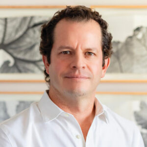 Un hombre de cabello castaño corto y rizado, vestido con una camisa blanca de corte impecable, posa en un espacio interior cuidadosamente diseñado. Al fondo, una selección de obras enmarcadas en blanco y negro —con representaciones artísticas de hojas a gran escala— realza la sofisticación del ambiente. Su mirada directa transmite seguridad, visión y clara mientras reflexiona sobre los desafíos actuales y las soluciones innovadoras que definen el desarrollo arquitectónico premium en la Baja. Se trata de un perfil orientado al inversionista o comprador exigente, que busca integrarse a proyectos inmobiliarios únicos donde diseño vanguardista, calidad constructiva y exclusividad crean nuevas oportunidades para maximizar el valor patrimonial.