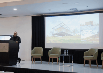 Un conferenciante especializado se presenta ante un selecto auditorio en un moderno salón de conferencias. En el escenario, el mobiliario minimalista -tres sillas exclusivas y una mesa con agua premium- subraya la atención al detalle y la calidad del entorno. La gran pantalla central proyecta información e imágenes sobre "Arquitectura del Nuevo Siglo", destacando innovaciones en diseño, sostenibilidad y lujo. Esta presentación está dirigida a inversores y compradores exigentes que buscan oportunidades únicas en desarrollos inmobiliarios de alto nivel, donde la excelencia arquitectónica se fusiona con el estilo de vida sofisticado.