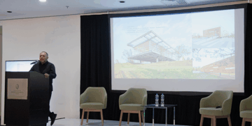 Un conferenciante especializado se presenta ante un selecto auditorio en un moderno salón de conferencias. En el escenario, el mobiliario minimalista -tres sillas exclusivas y una mesa con agua premium- subraya la atención al detalle y la calidad del entorno. La gran pantalla central proyecta información e imágenes sobre "Arquitectura del Nuevo Siglo", destacando innovaciones en diseño, sostenibilidad y lujo. Esta presentación está dirigida a inversores y compradores exigentes que buscan oportunidades únicas en desarrollos inmobiliarios de alto nivel, donde la excelencia arquitectónica se fusiona con el estilo de vida sofisticado.